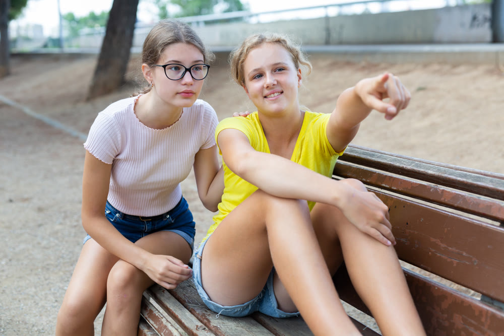 Two girlfriend sitting on bench and points to something