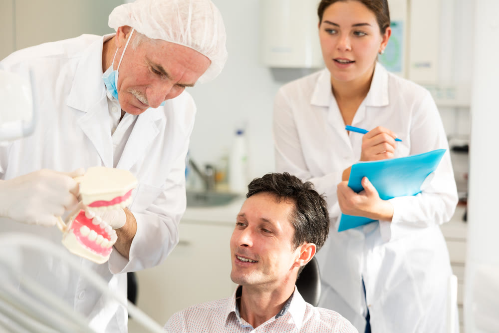 Senior man dentist and girl nurse communicates with male patient, shows model of jaws