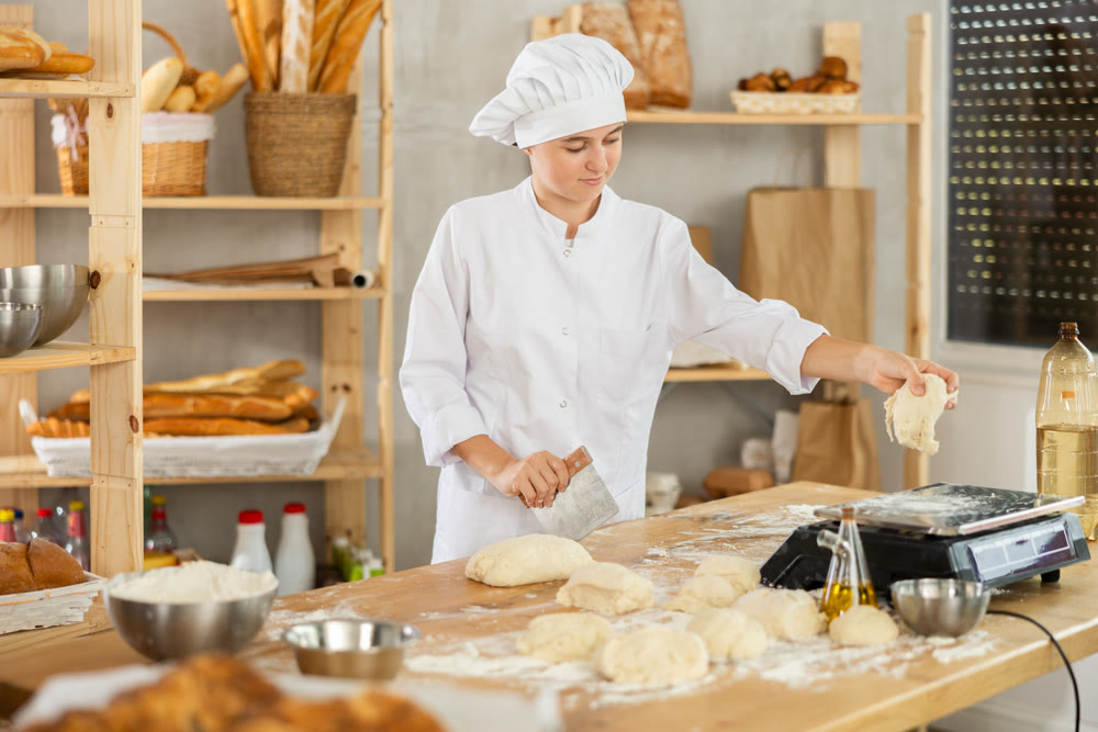 Baker teen girl cuts small pieces of dough from large lump and puts them on scales.