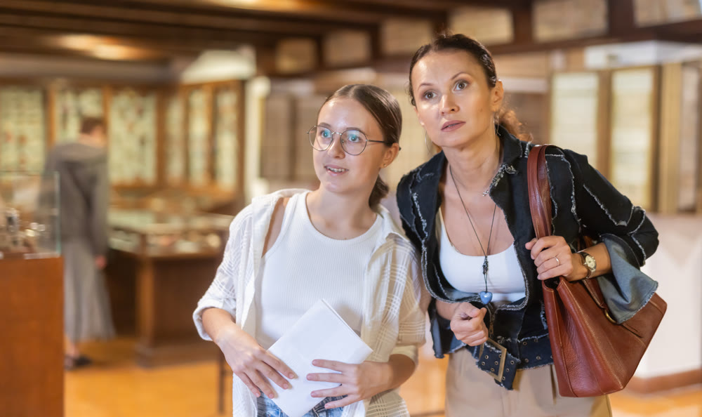 Young woman with teen daughter walk around museum, view antique appliances in museum hall