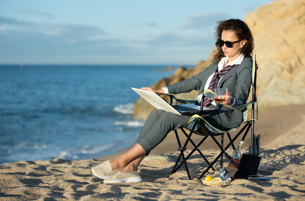 Businesswoman sits on a beach chair with a laptop and whiskey