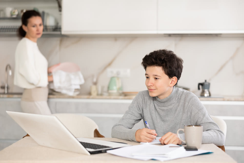Teen son doing school homework while mom washes dishes in kitchen