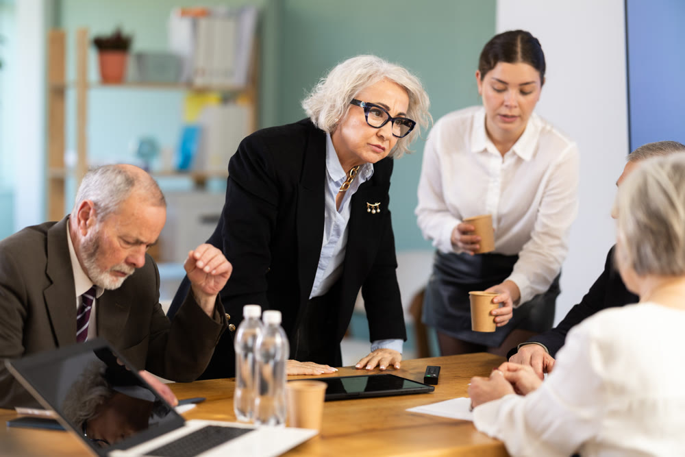Elderly woman standing and scolding sad senior colleagues at meeting table