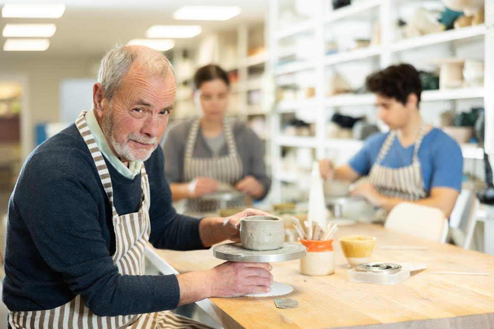 Mature man at table molds cup from wet clay in pottery workshop