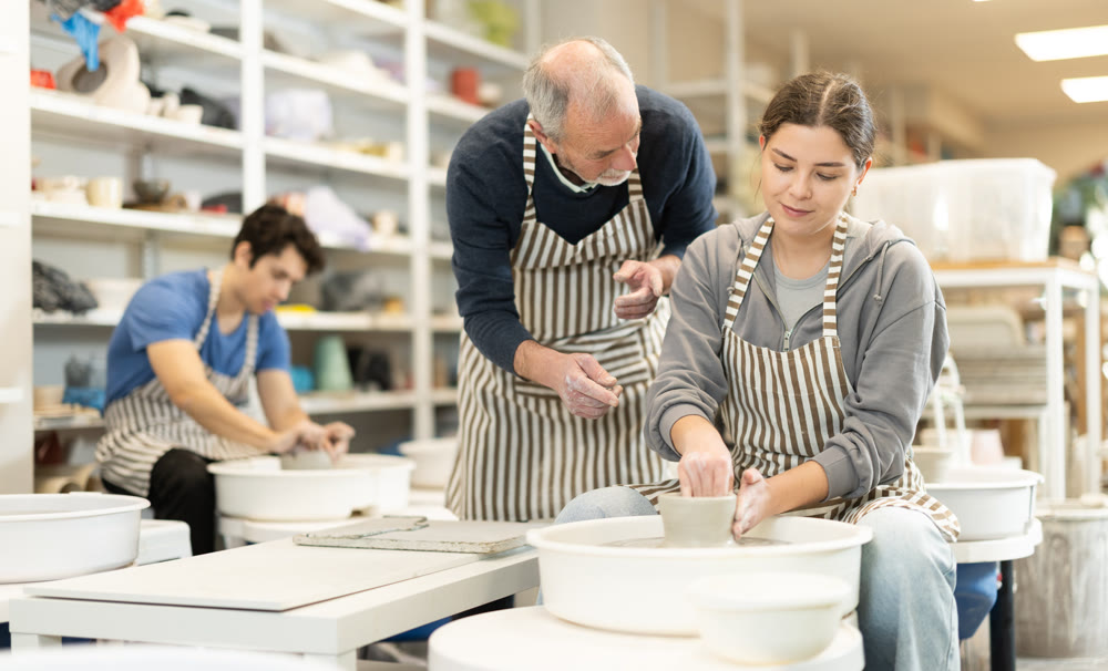 Mature man help to girl working on potters wheel in pottery workshop