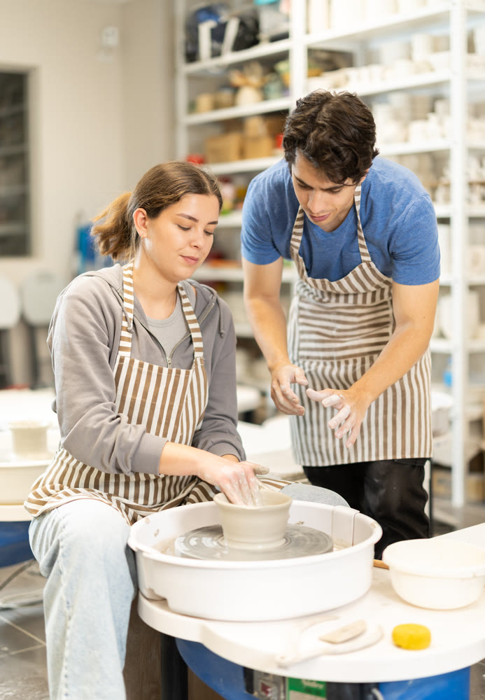 Young man help to girl working on potters wheel in pottery workshop