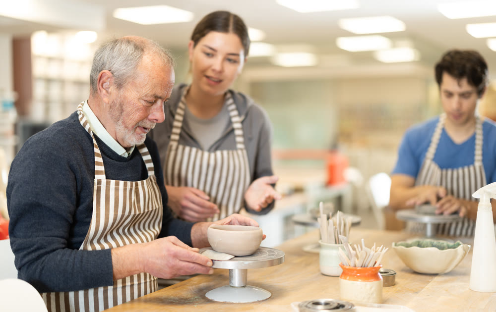 Woman help mature man sculpting ceramic product from raw clay in workshop