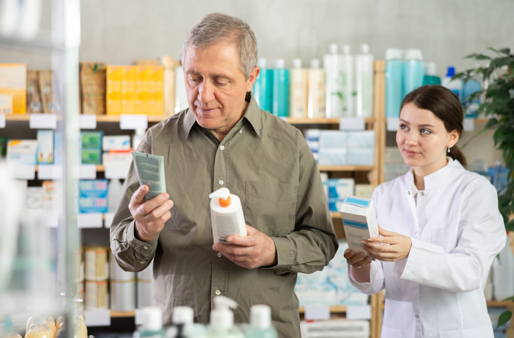 Senior man buys cream at drug store, girl pharmacist in background