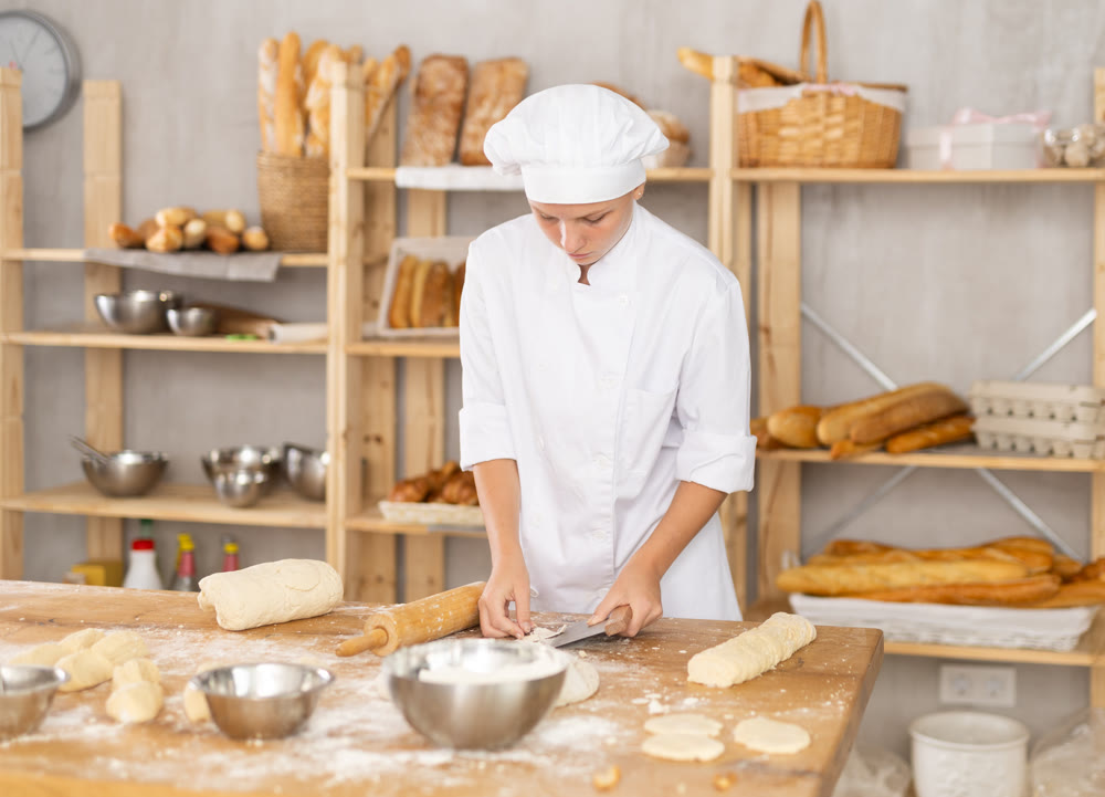 Young female baker cutting dough with scraper