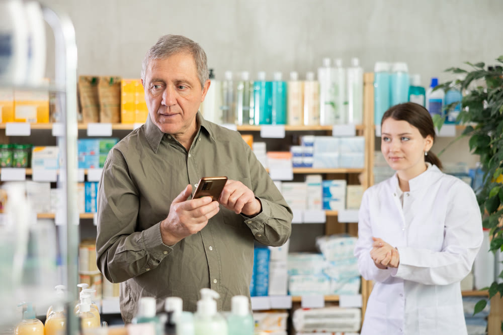 Senior man with phone is shopping in pharmacy. Girl pharmacist offer help to customer