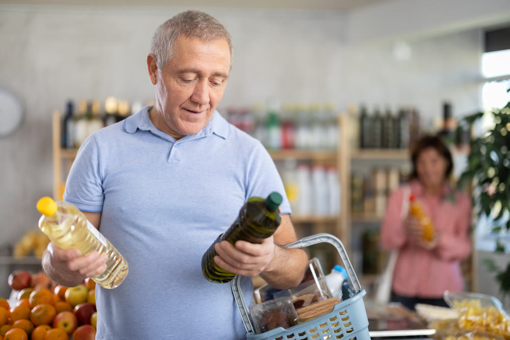 Mature man buyer looks thoughtfully at bottles of vegetable oil, woman client in background