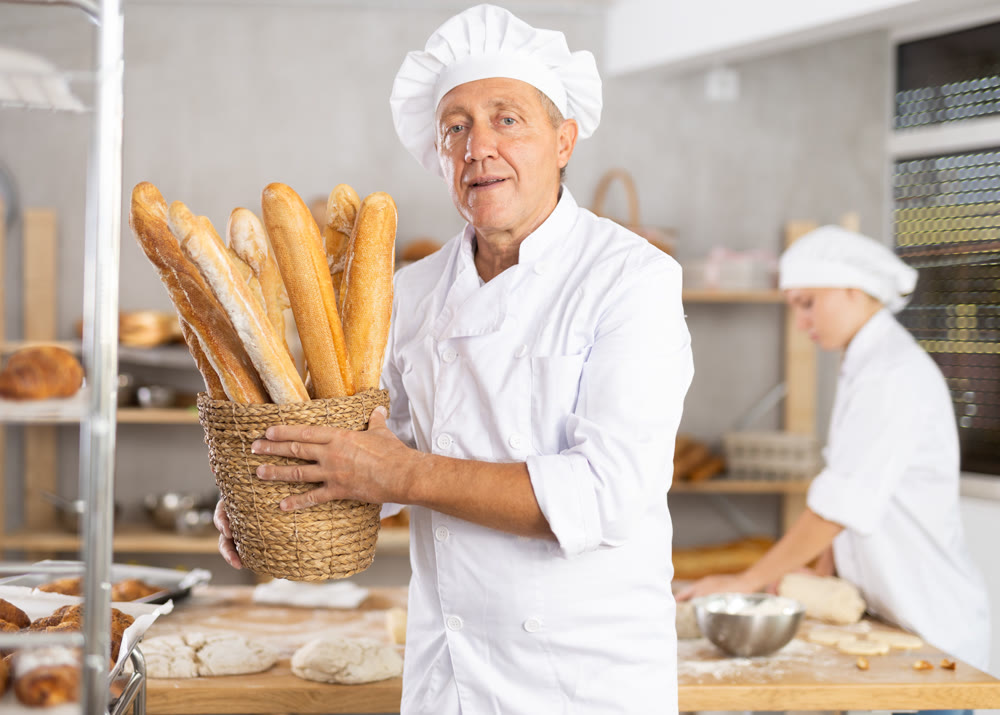 Portrait of elderly baker with bread and baguettes in hands in kitchen