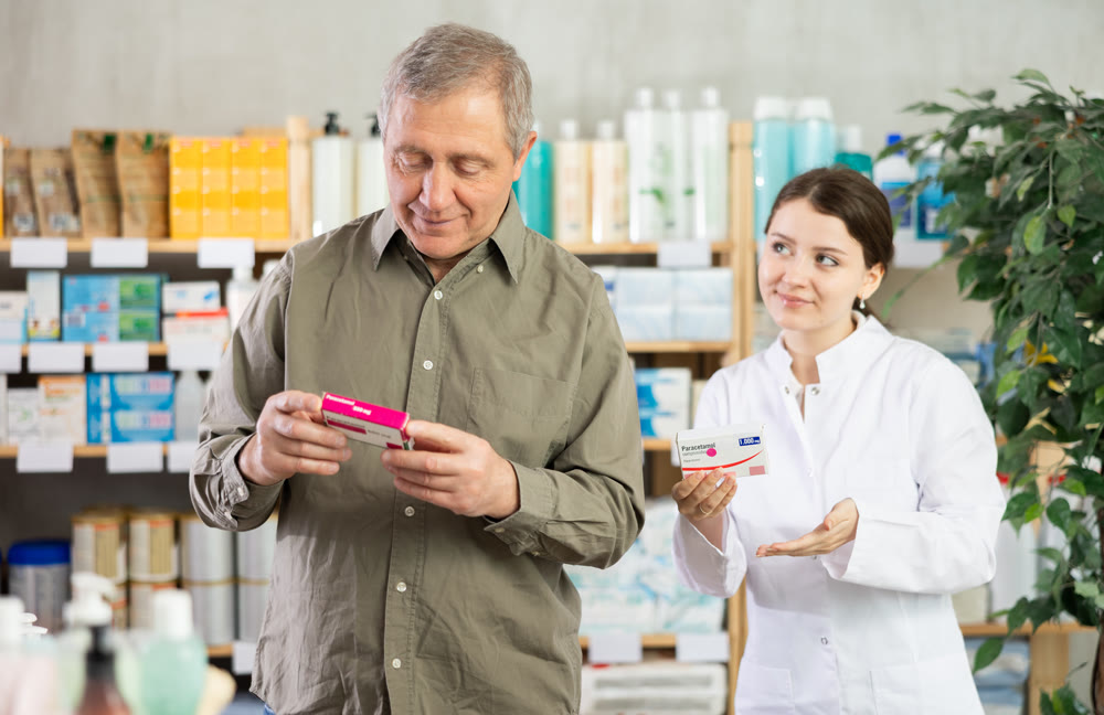 Senior man buys pills at pharmacy. Girl pharmacist in background