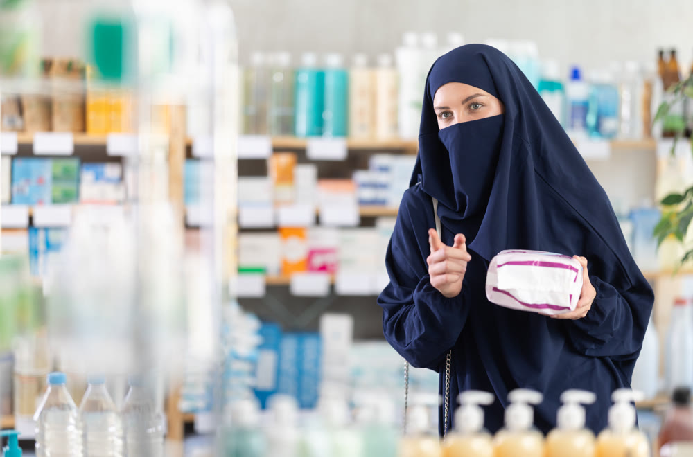 Young woman in Muslim attire choosing feminine pads in pharmacy