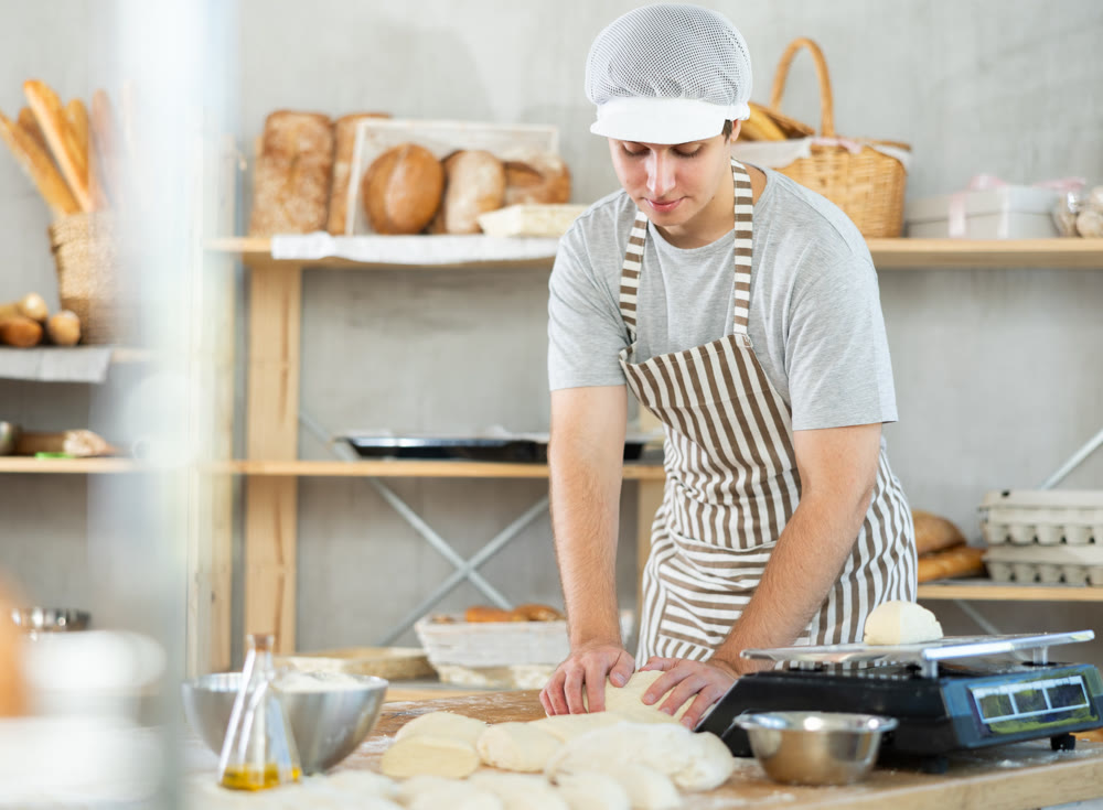 Young positive male baker in apron kneading and forming dough on table, process of preparing of bread in kitchen