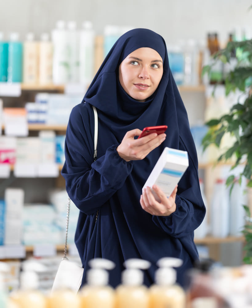 Young Muslim woman scanning QR-code on cream in drugstore