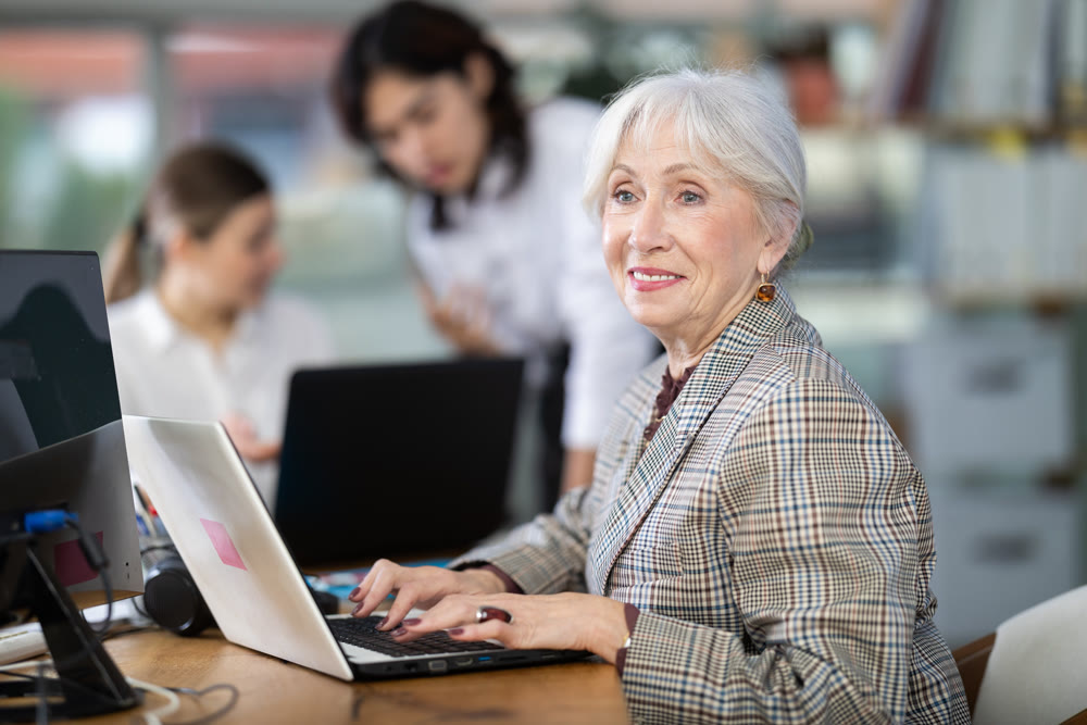In office, senior woman is working on laptop as part of group of colleagues.