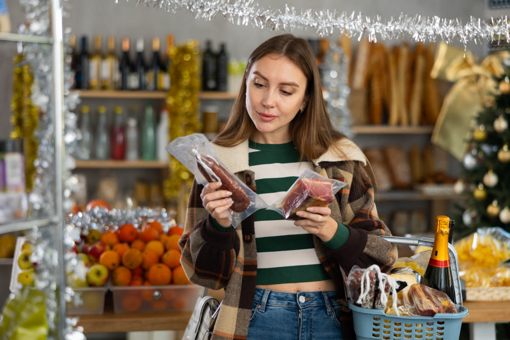 Woman with grocery cart choosing dried sausage and jamon for Christmas or New Year celebration in produce section of supermarket