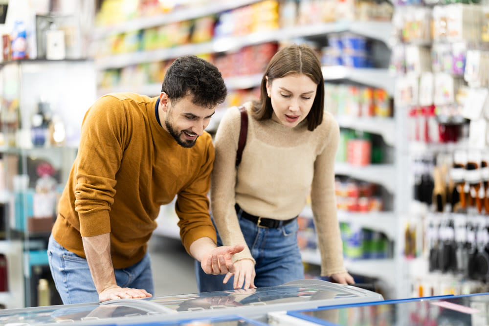 Positive young couple choosing cooled food product standing at fridge in grocery store