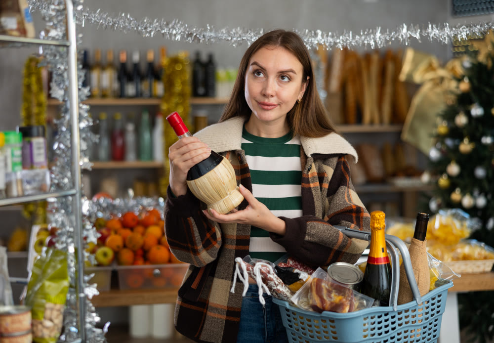 Beautiful attentive young girl buyer with shopping cart or basket choosing wine for christmas table at grocery