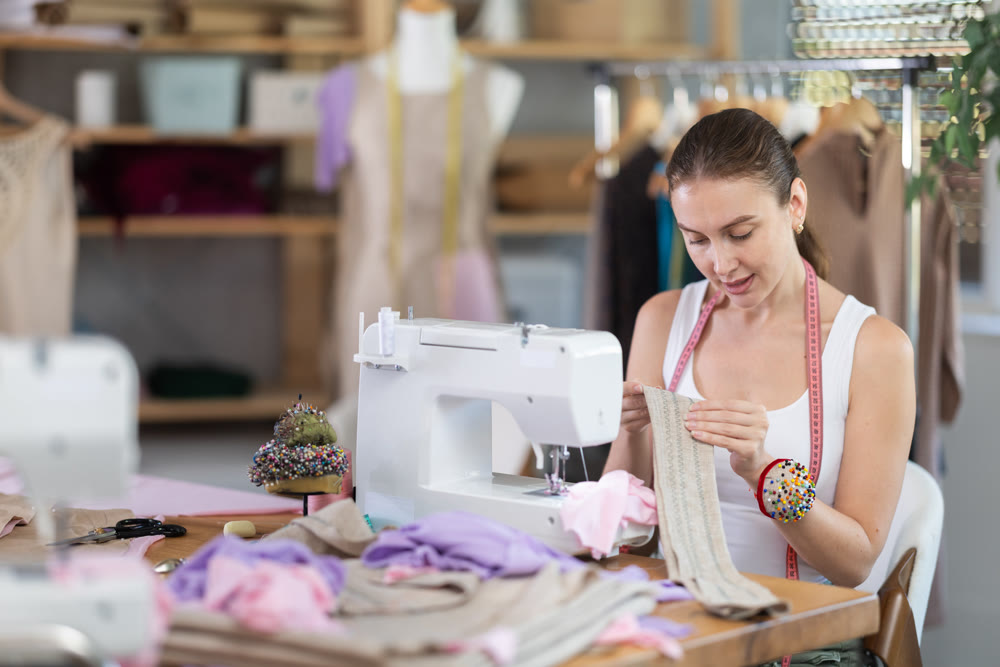 Glad young dressmaker viewing dress detail sitting at sewing-machine