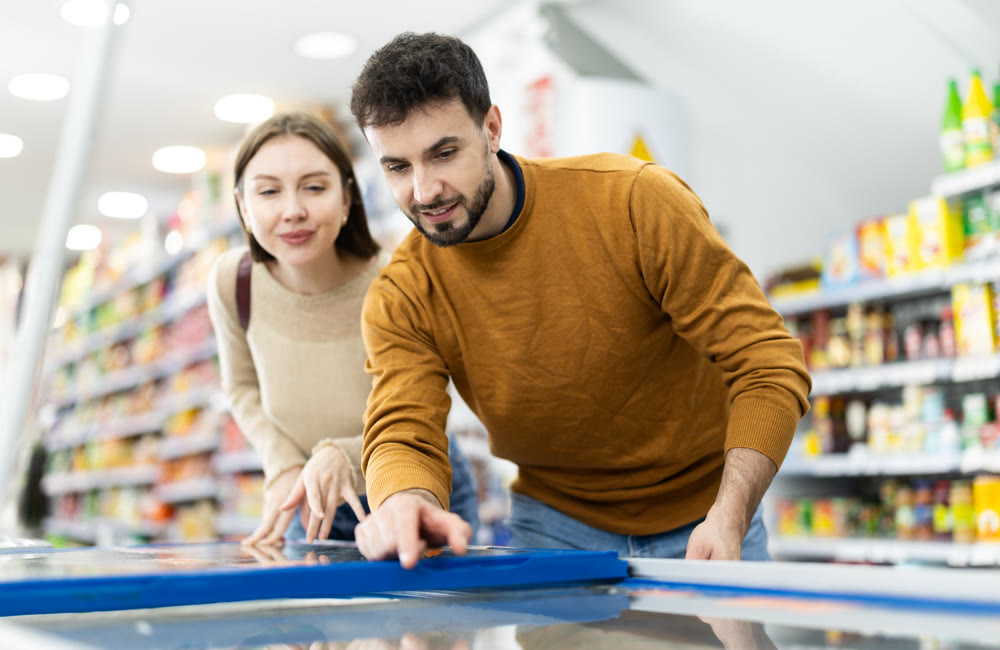 Positive young couple choosing cooled food product standing at fridge in grocery store