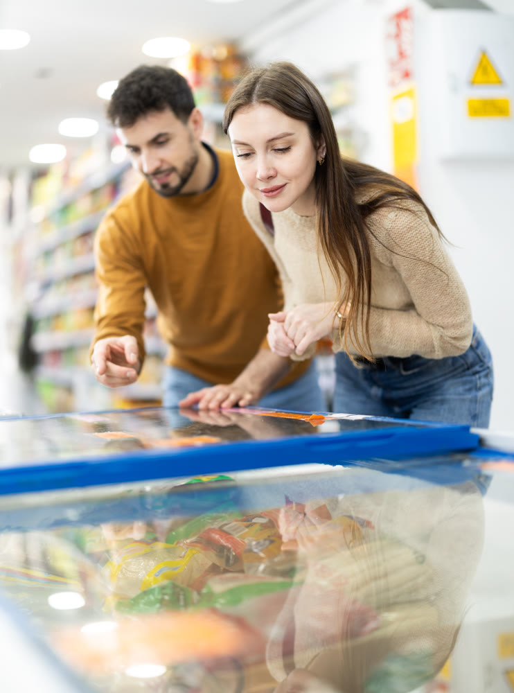 Positive young couple choosing cooled food product standing at fridge in grocery store