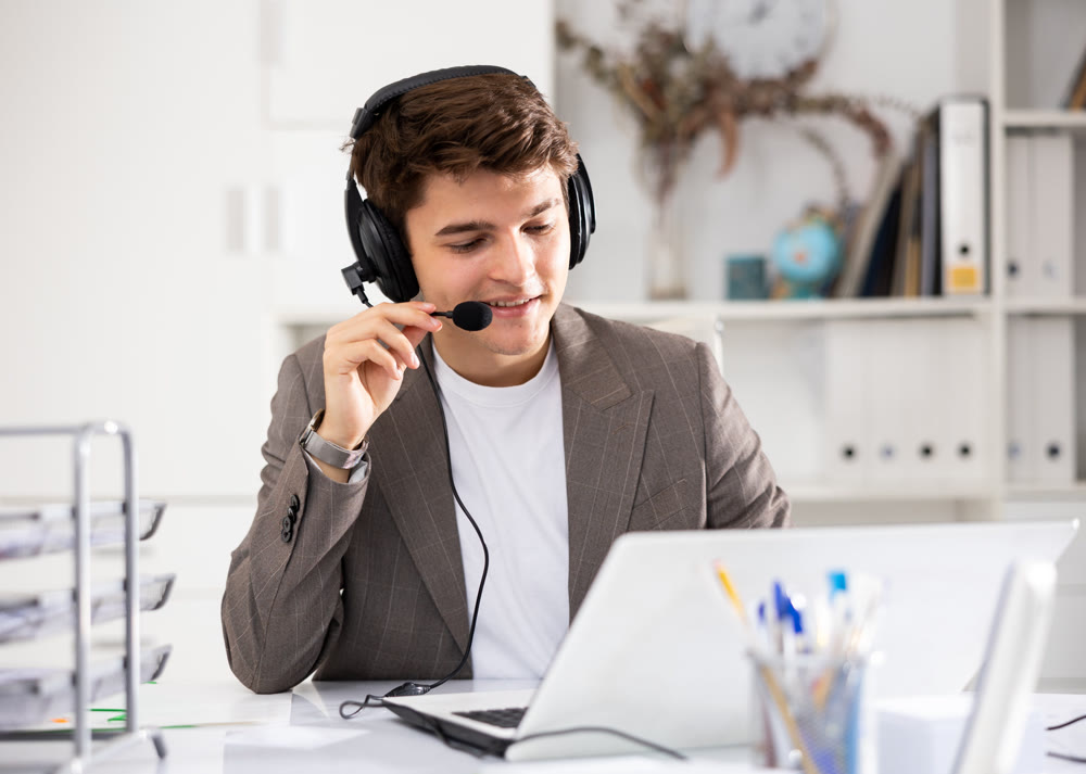 Positive businessman with headphones and microphone having video call on laptop in office