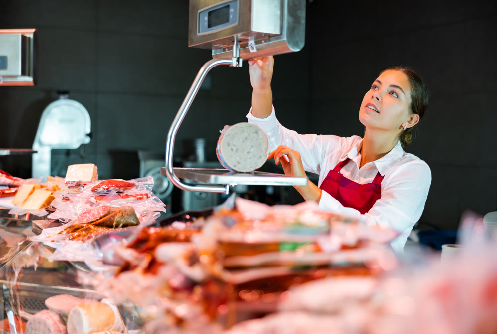 Young female seller of grocery store weighing mortadella on scales