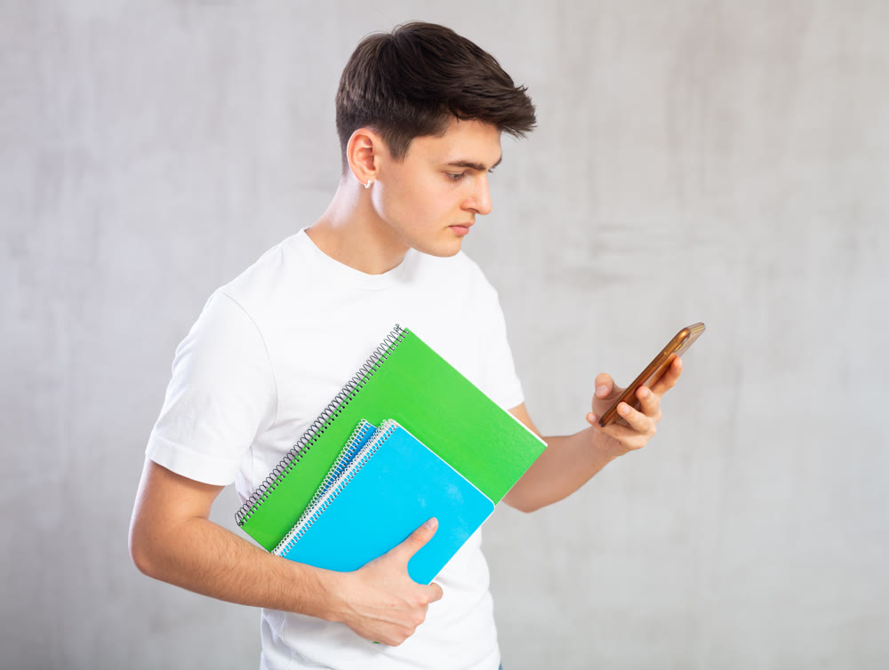 Attentive young student with workbooks in hands looking at mobile phone