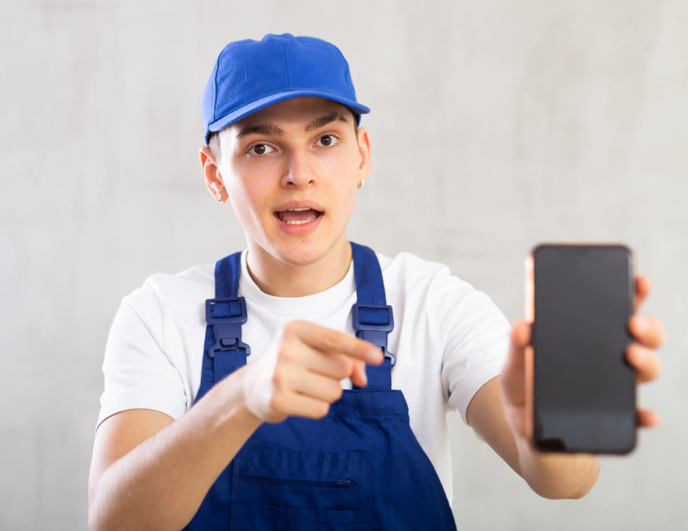 Man in jumpsuit and cap shows dark empty mobile phone screen and points at device with his hand