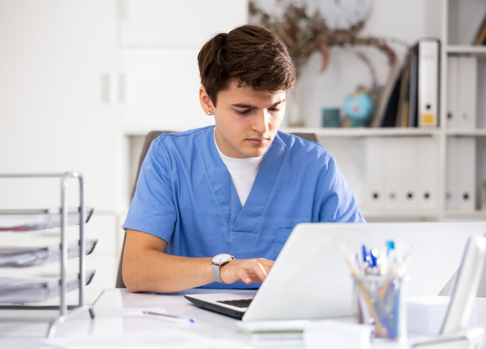 Positive man doctor therapist sitting in office at workplace and working at laptop