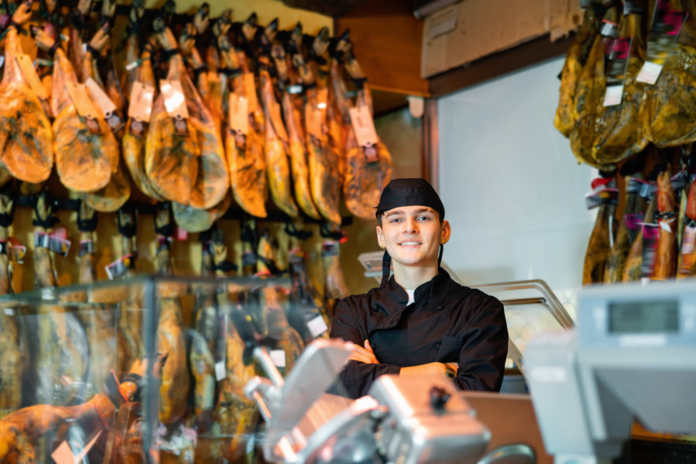 Young butcher shop salesman in black uniform standing with crossed arms near rack with hanging jamon legs