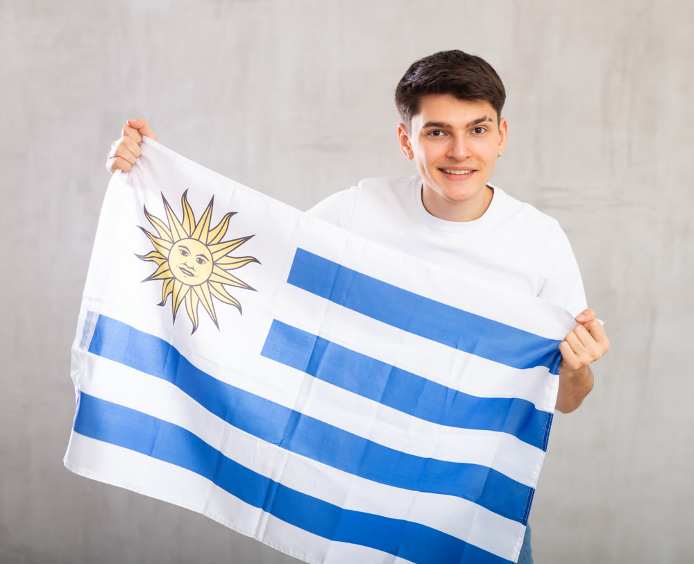 Happy young man holding flag of Uruguay against unicoloured background