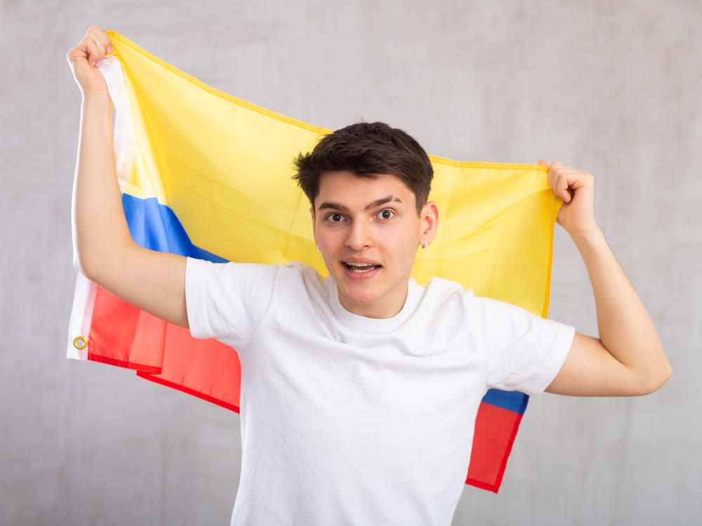 Man in holds unfurled flag of Colombia in hands raised above head against gray wall