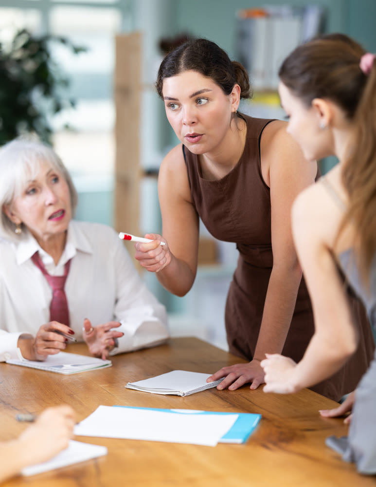 Concentrated businesswoman standing with pen in hands near table where sitting female colleagues different ages and talking in corporate seminar in office