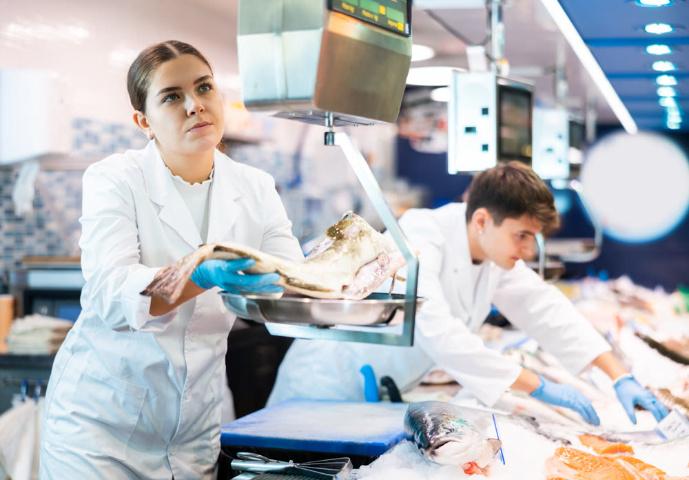 Female seller in fish department of supermarket weighs atlantic cod fish on scale