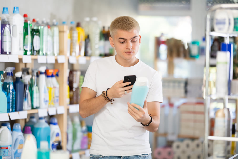Positive young man buyer in shop holding and choosing shampoo or shower gel, scan QR code on label