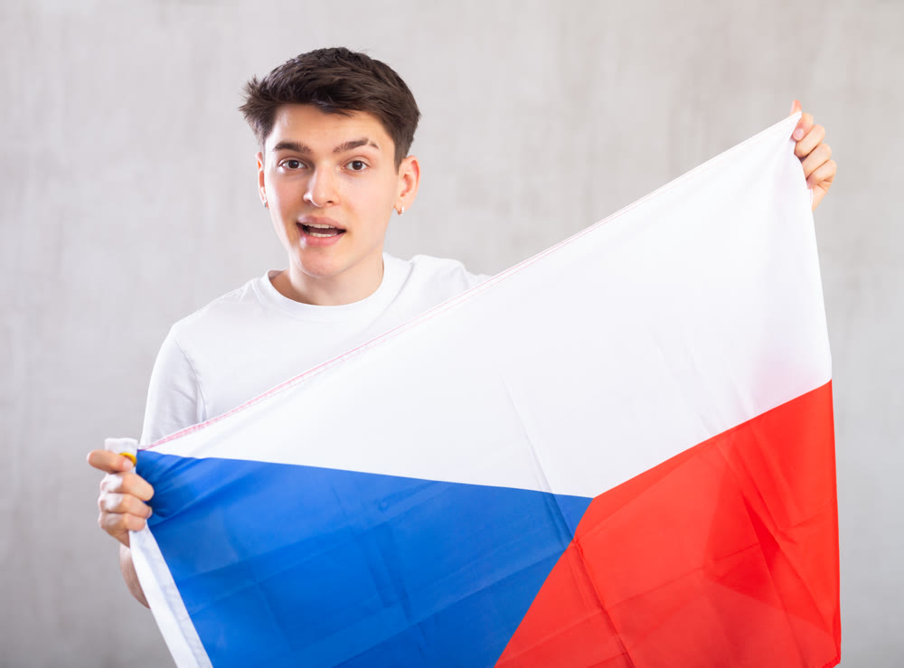 Man in casual clothes holds unfurled flag of Czechia in hands raised above head against gray wall