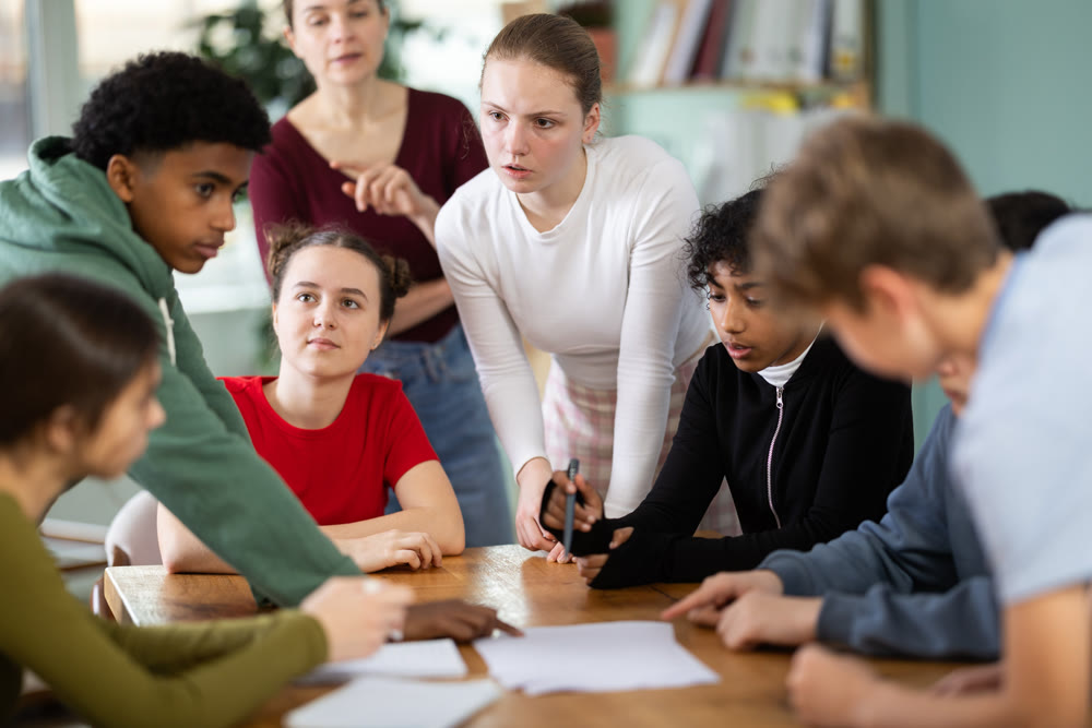 Teenage students solving problem on paper while teacher watches behind