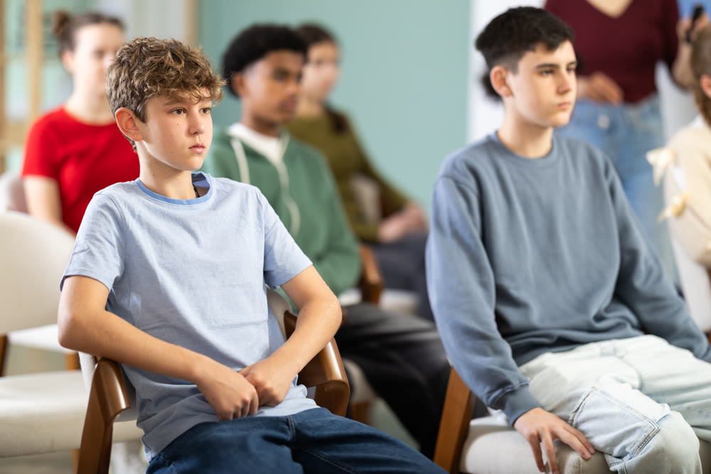 Teenage boy listening to teacher while sitting with classmates in classroom