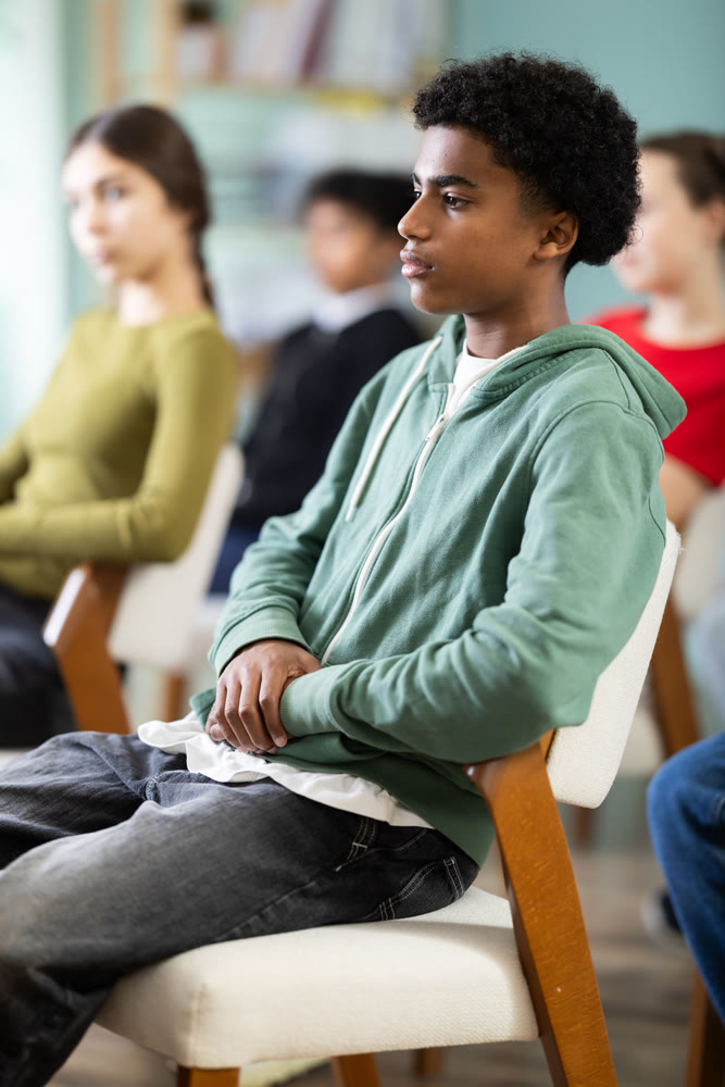 Teenage boy listening to teacher while sitting with classmates in classroom