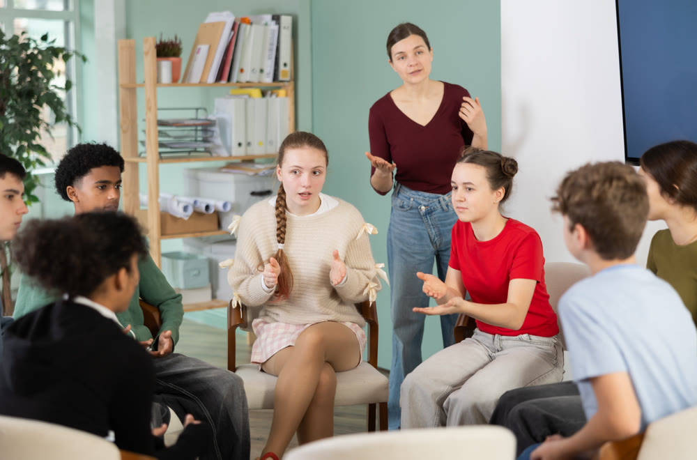 Teenage students sitting in circle and talking under supervision of teacher
