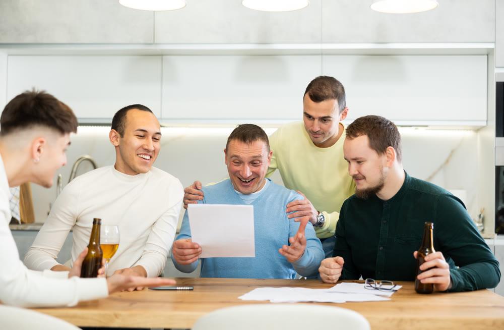 Happy friends drinking beer after solving their financial problems while sitting together at home cooking table
