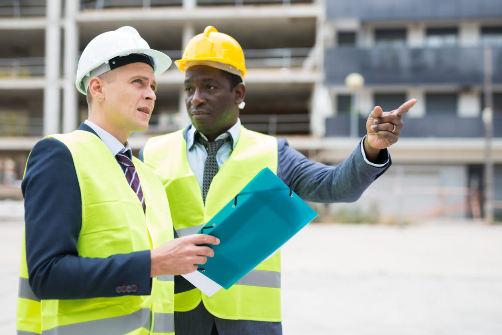 African-american and European men standing on construction site and talking. One of them holding folder with documentation, another one showing something with finger gesture.