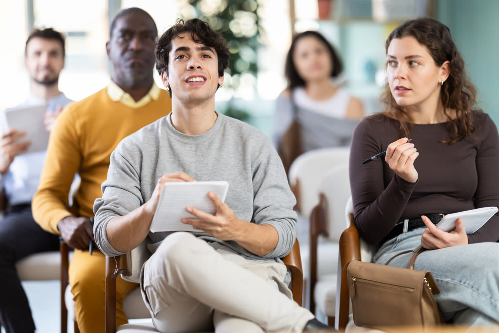 During lecture meeting, young man pupil listen to teacher, write down summary.