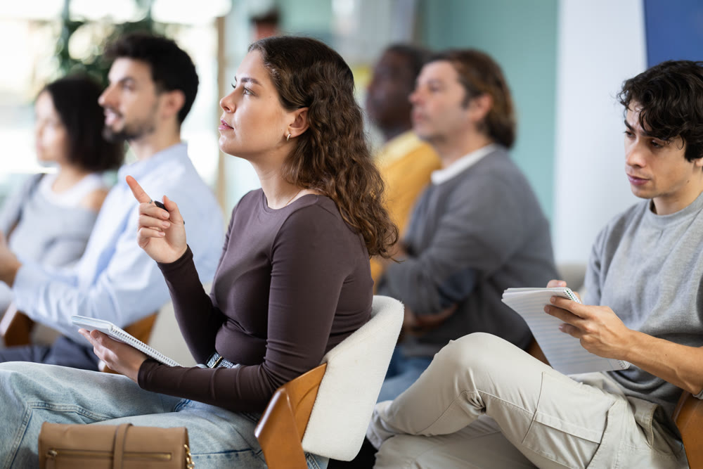 During lecture meeting, young woman pupil listen to teacher, write down summary.