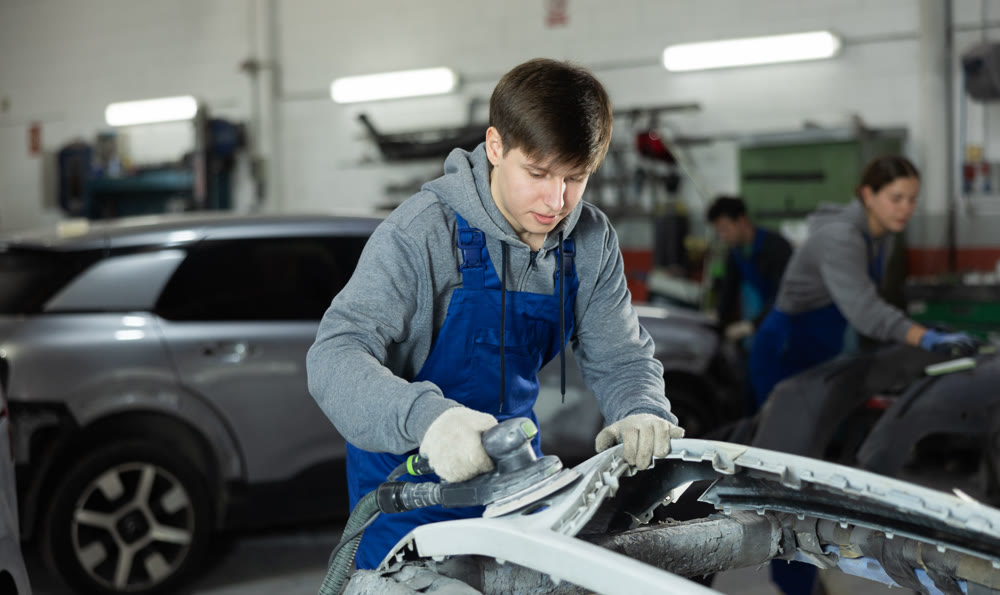 Young man auto mechanic polishes body parts by car buffer polisher machine tool
