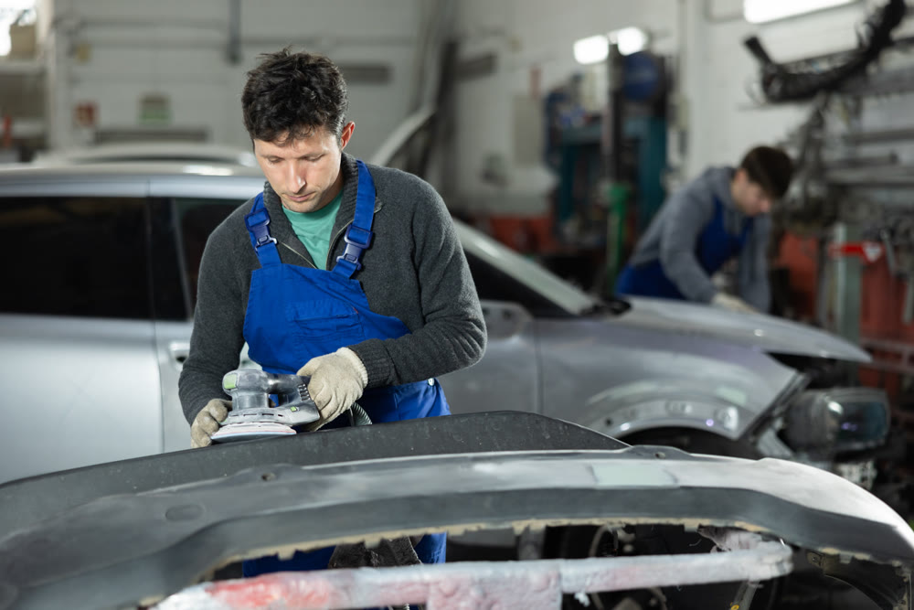 Male mechanic in uniform polishes bumper of car with machine in car service station