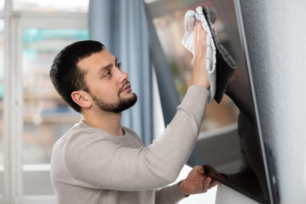 Young man rubbing screen of wall-mounted TV in apartment